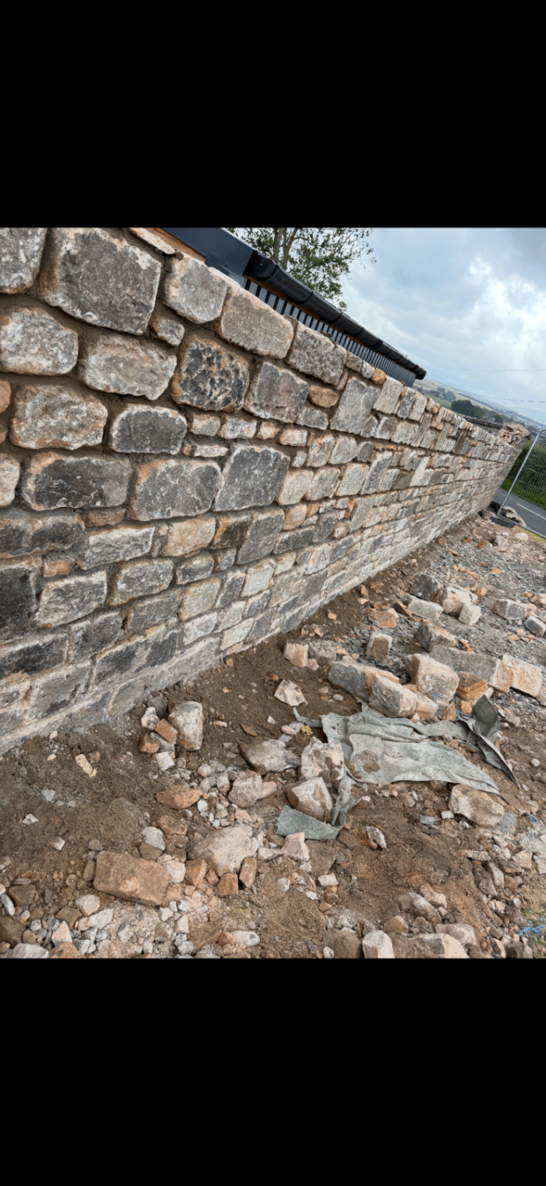 Long hand-built natural stone boundary wall under construction with rubble and prepared ground in the foreground.