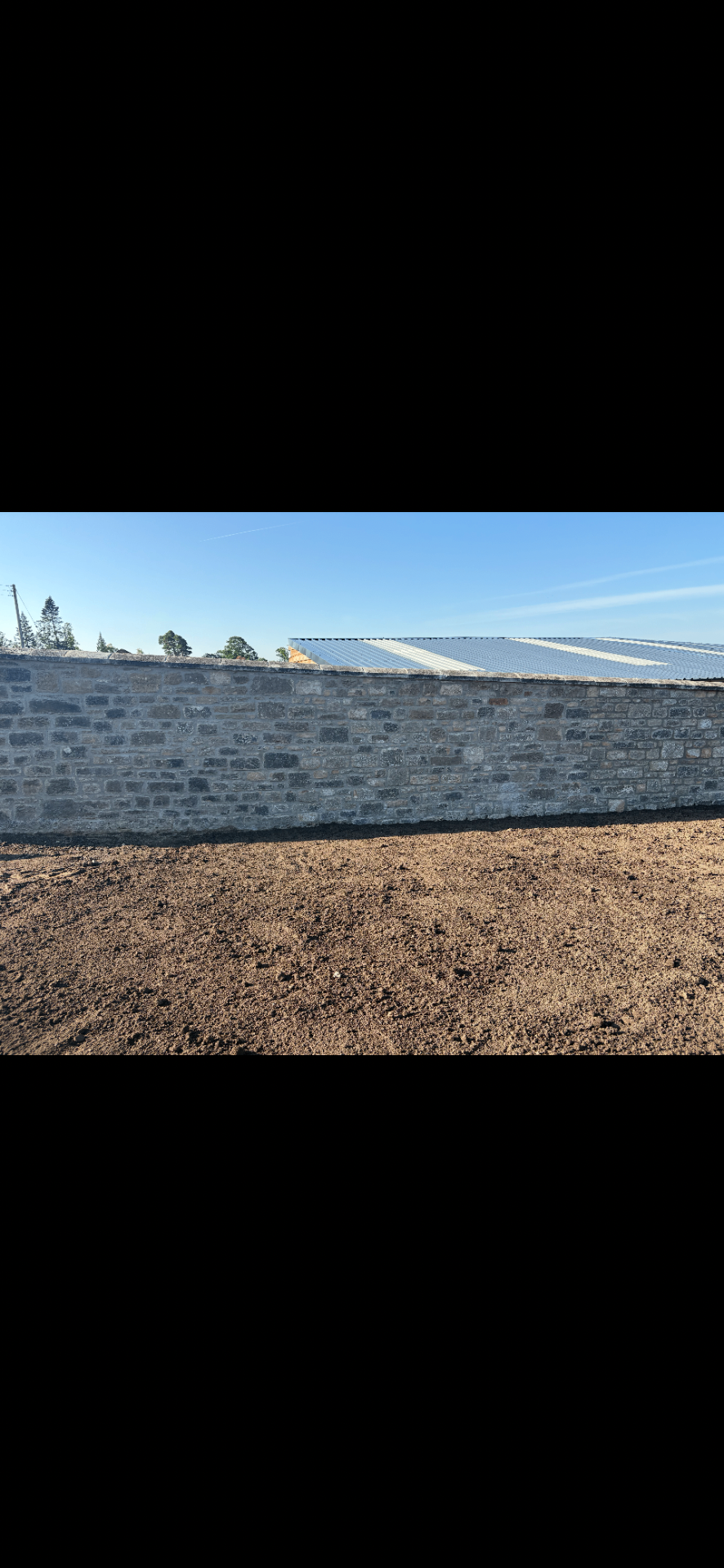 Long finished natural stone boundary wall with a level prepared foreground and agricultural buildings behind.