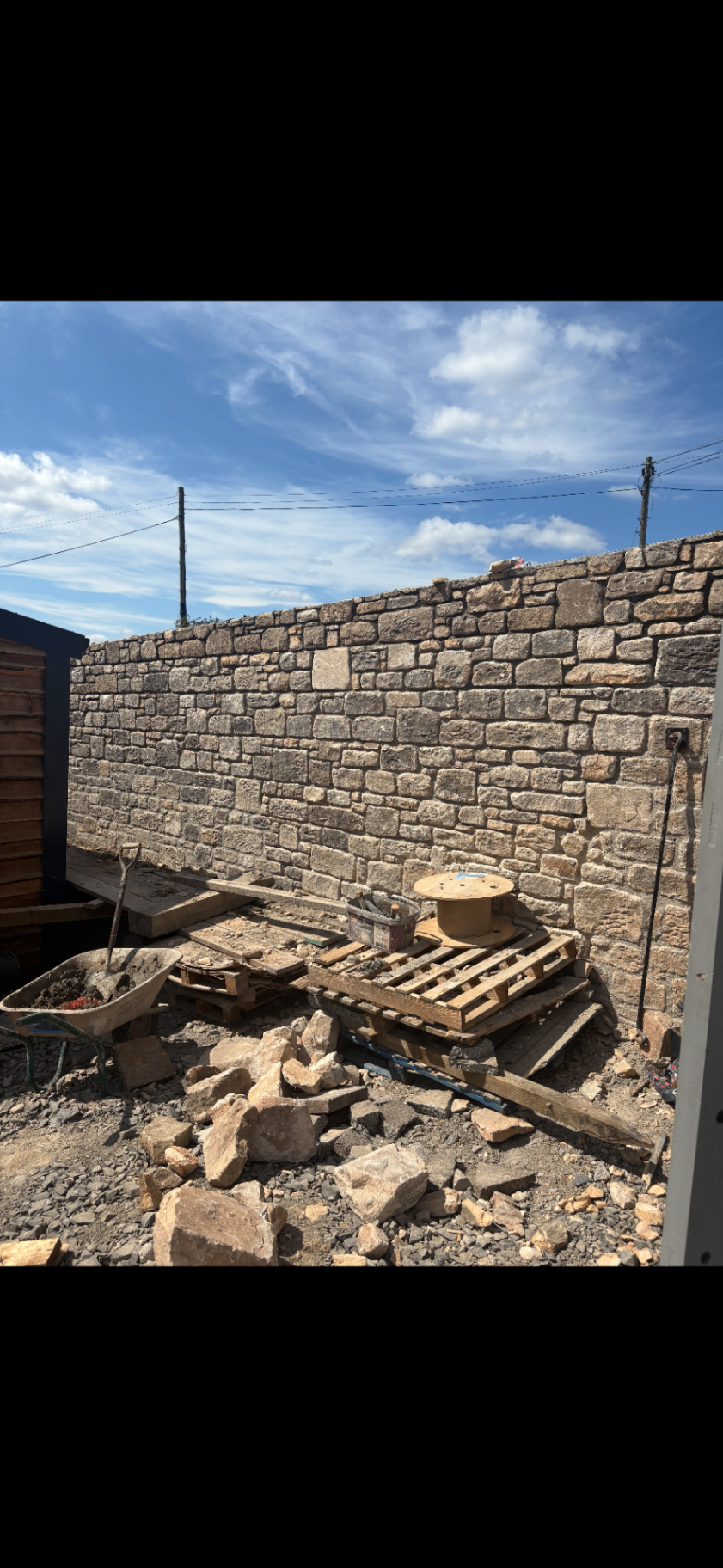 Natural stone boundary wall under construction with loose stone, tools and pallet materials in the foreground.