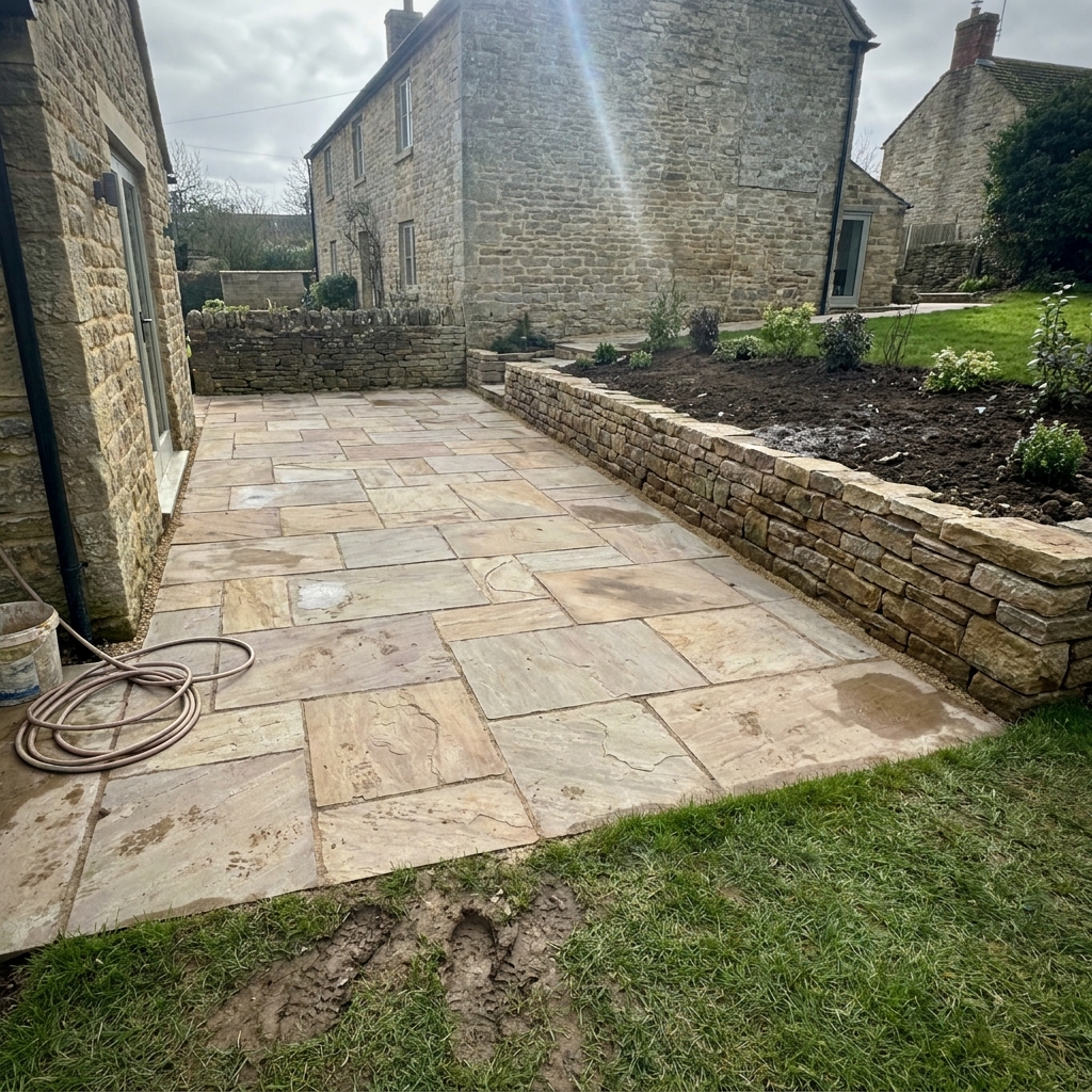 Newly laid Indian sandstone patio with a hand-built dry stone retaining wall and freshly planted borders beside a traditional stone cottage.